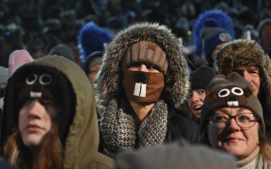 A crowd dressed in warm clothing, some decorated with groundhogs, waits Punxsutawney Phil.