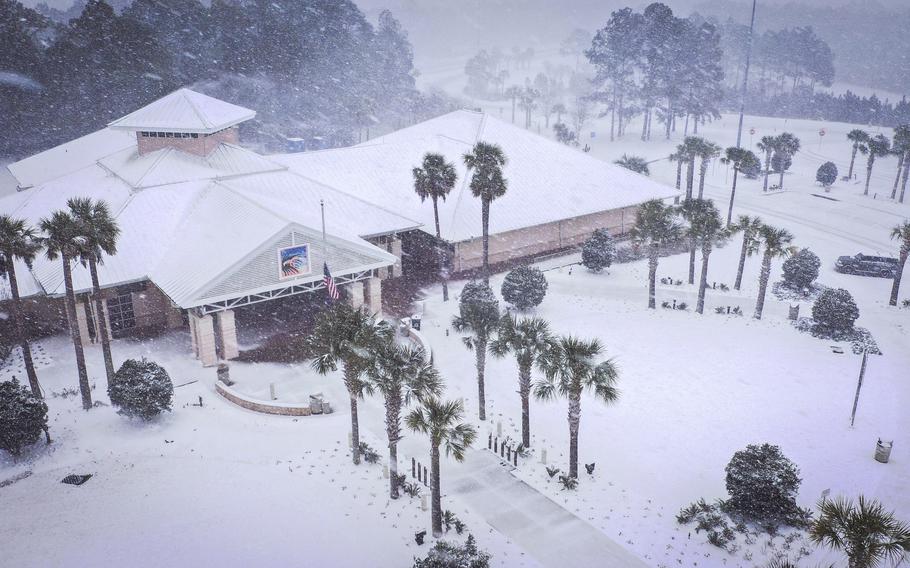 An aerial photo of a large one-story building with a long walkway and palm trees, all covered in snow.