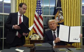 Donald Trump sitting at his desk in the Oval Office holds up an executive order.