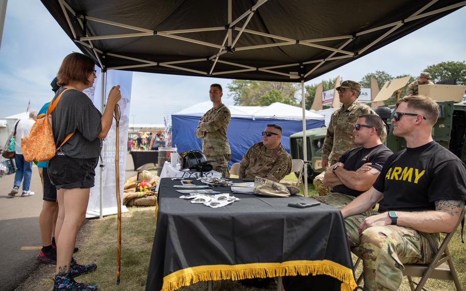 Soldier sit at a table with informational pamphlets and speak with young people at a recruitment booth.