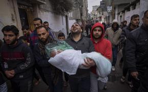 A group of Palestinian men walk down a street, with one, his face twisted in anguish, carrying the plastic and blanket wrapped body of a young girl.