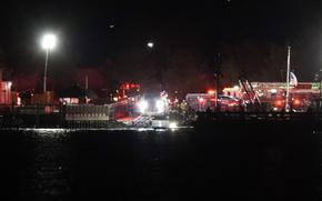 Emergency vehicles with flashing lights, seen from a distance, park along a darkened river front.