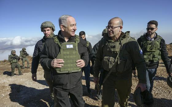 Israel’s Prime Minister Benjamin Netanyahu on the left and with his hands on an armored vest, smiles while looking at the person on the right.