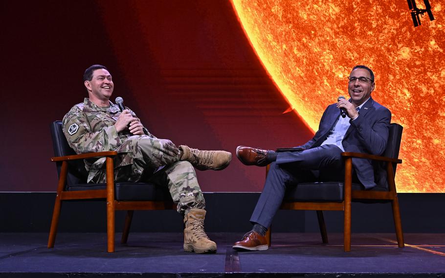 Two men sit close to each other on a stage, one on the left in a uniform and the other on the right in a suit, each with their legs crossed.