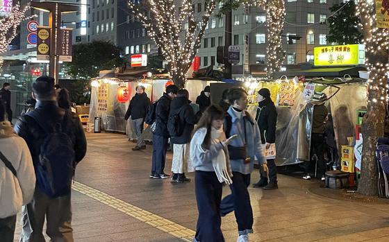 Japanese pedestrians walk on a street in front of lighted merchandise stalls and trees festooned with white lights.