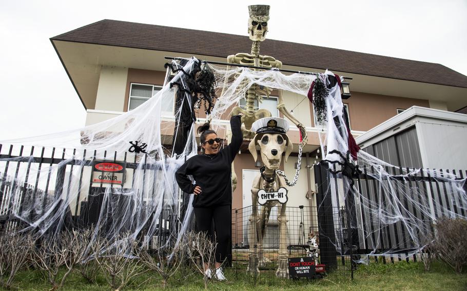 A woman in a black sweatsuit poses in front of a house next to Halloween decorations of a large skeleton and a skeletal dog.