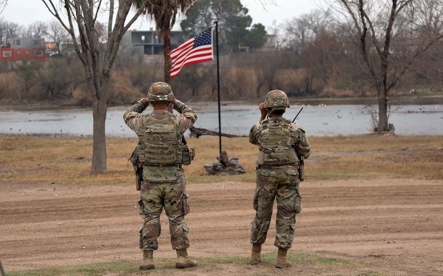 Two soldiers looking at a river with the American flag flying between them.