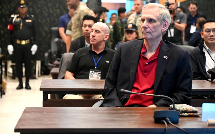 An American ambassador in a dark sports coat and red shirt sits at a table and listens to a speaker during a conference, with other attendees seated at tables behind him.