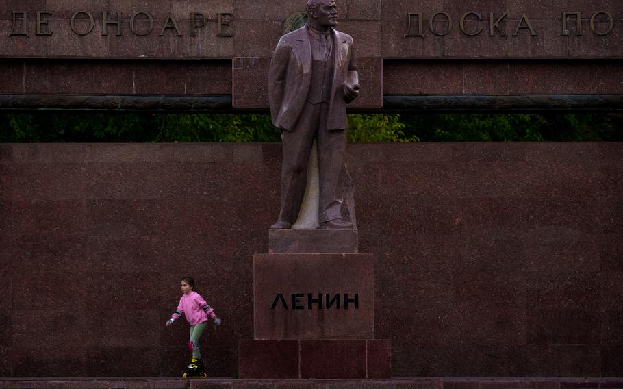 A large statue of Lenin in the center of the photo with a young girl skating next to it.