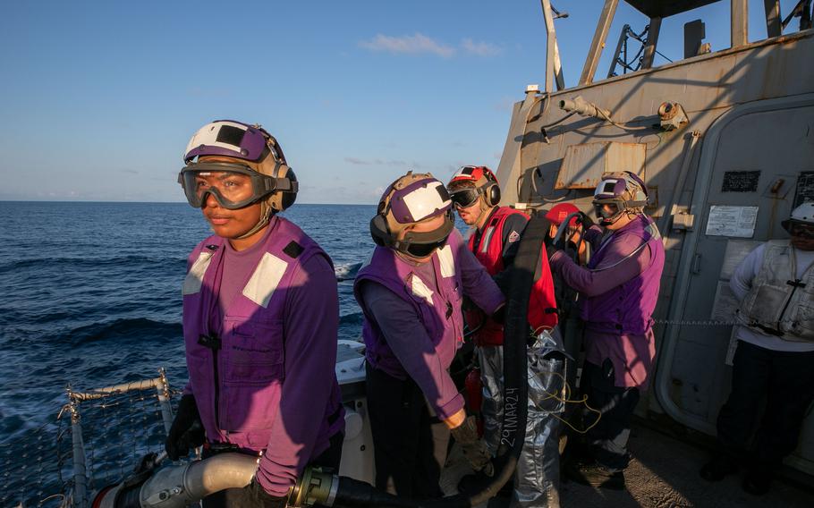 Sailors wearing goggles and dressed in purple jackets and black gloves hold a tube on a navy ship.