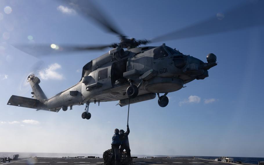 A MH-60R Sea Hawk helicopter hovers above a pair of sailors attaching a cargo hook.