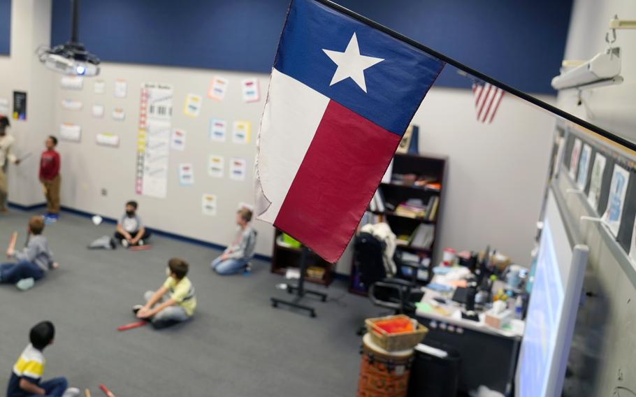 Elementary school children sit on a carpet beneath a Texas state flag in a classroom.