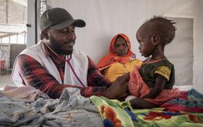 A doctor smiles while treating a young child, with a woman in the background watching the child.