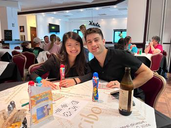 Man and woman sitting at a table, looking at the camera, playing BINGO. The table is covered with BINGO cards and BINGO ink dabbers.