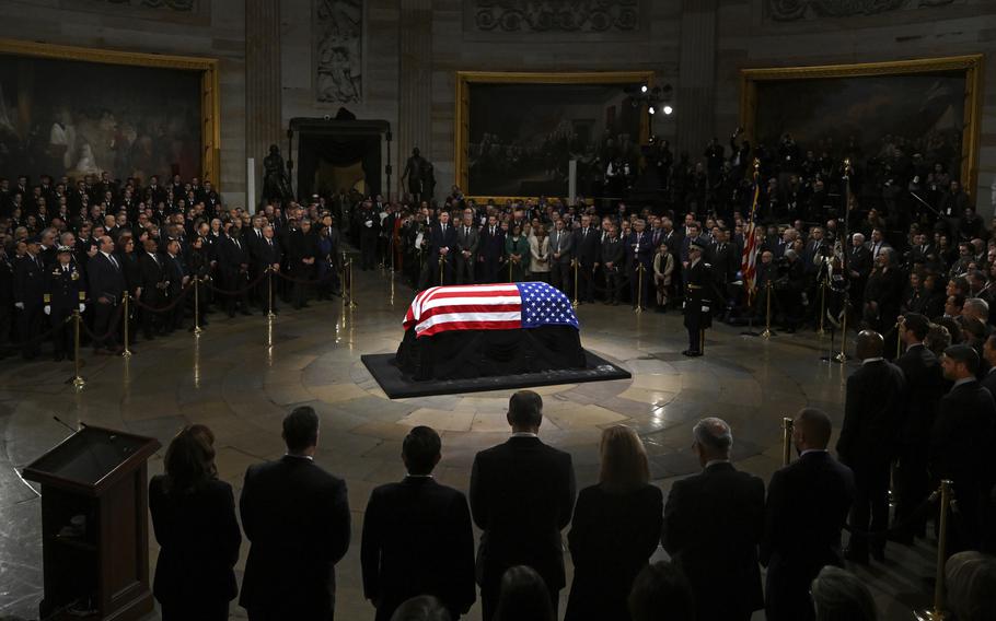 A casket draped with a U.S. flag sits on a pedastal in the atrium of the Capitol building as mourners stand in a circle around it.