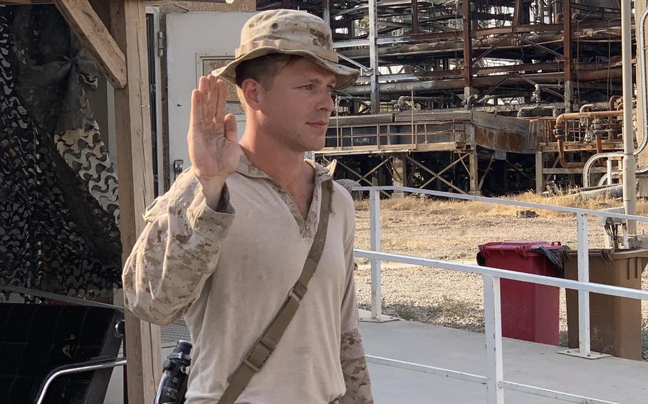 A Marine in camouflage uniform and boonie hat stands with his right hand raised.