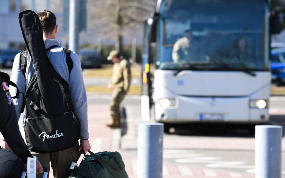 A man with a guitar strapped to his back and other luggage stands at the airport with a bus stopped nearby.