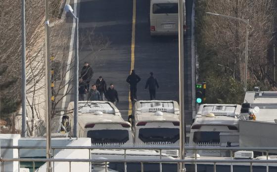 A security fence is seen barricaded by buses, with security personnel walking on the road in front of the buses.