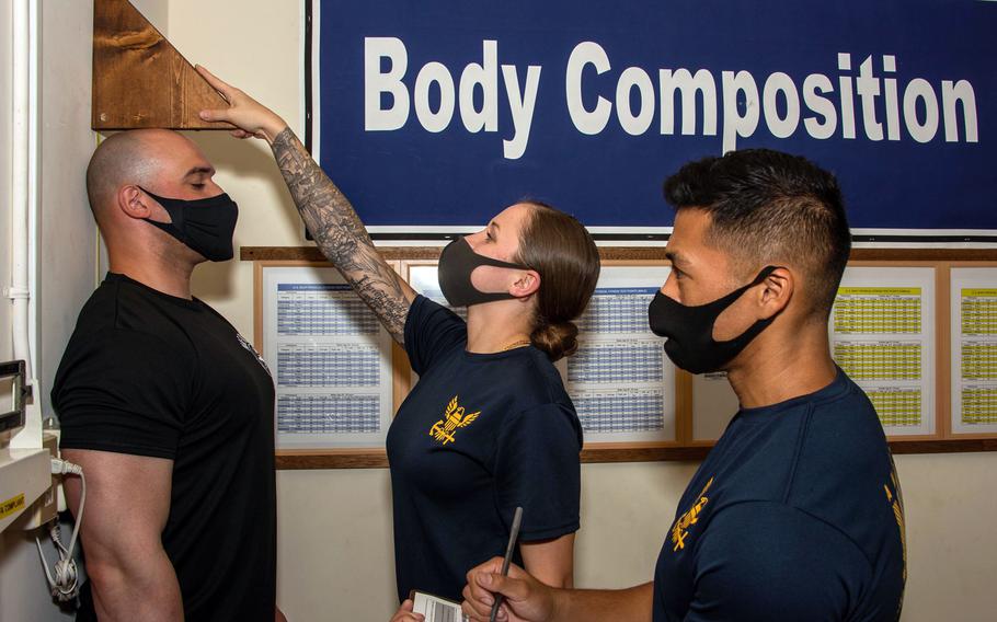 In front of a sign reading “body composition,” a sailor wearing a mask measures the height of another sailor as a third sailor looks on. 