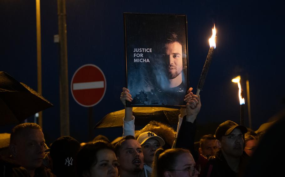 A group of protesters gather in the dark outside Spangdahlem Air Base. Two hold torches, one holds a sign that reads “Justice for Micha.”