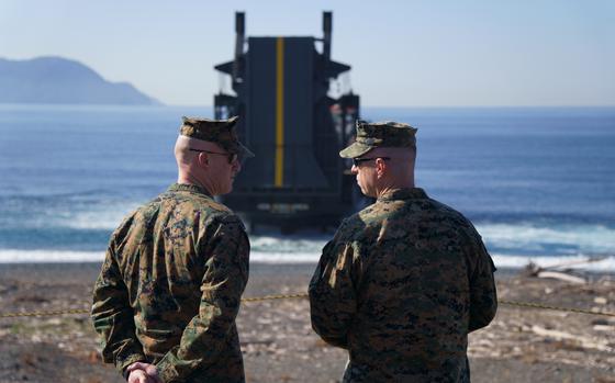 Two Marines in camouflage, seen from behind, speak to each other on a beach with a ship in the shallows of a bay in the background.