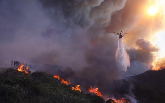 A helicopter on the right side of the image drops water over burning hills.