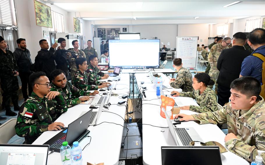 American and Thai soldiers in camouflage uniforms sit at cimputers on opposite sides of a long table with a large screen at one end.