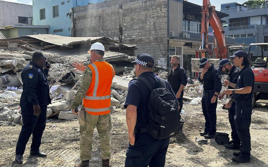 A ring of rescue workers, mostly in dark blue or black, stand in front of a collapsed building.