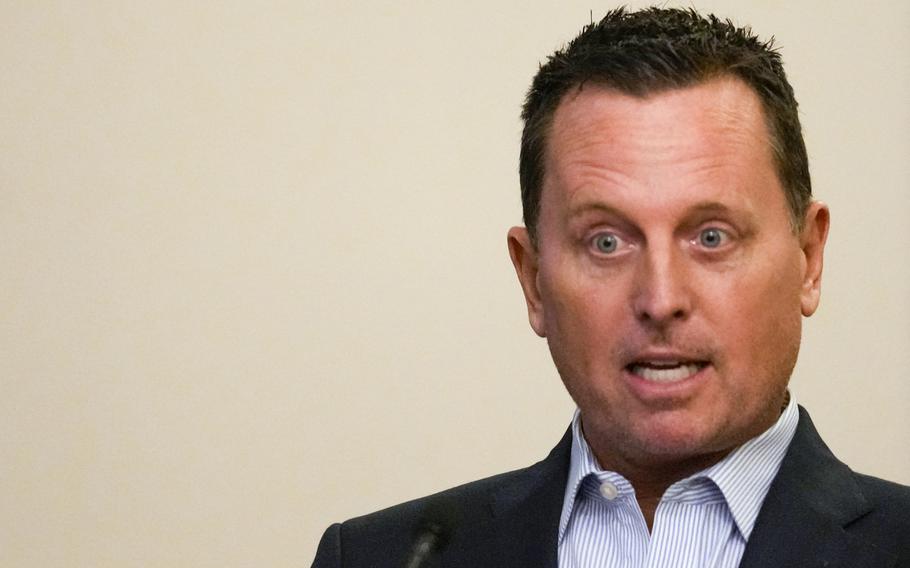 A man in a black suit and striped blue button down shirt speaks in front of a cream colored wall.