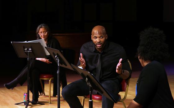 Three actors sit in front of music stands with scripts on them.