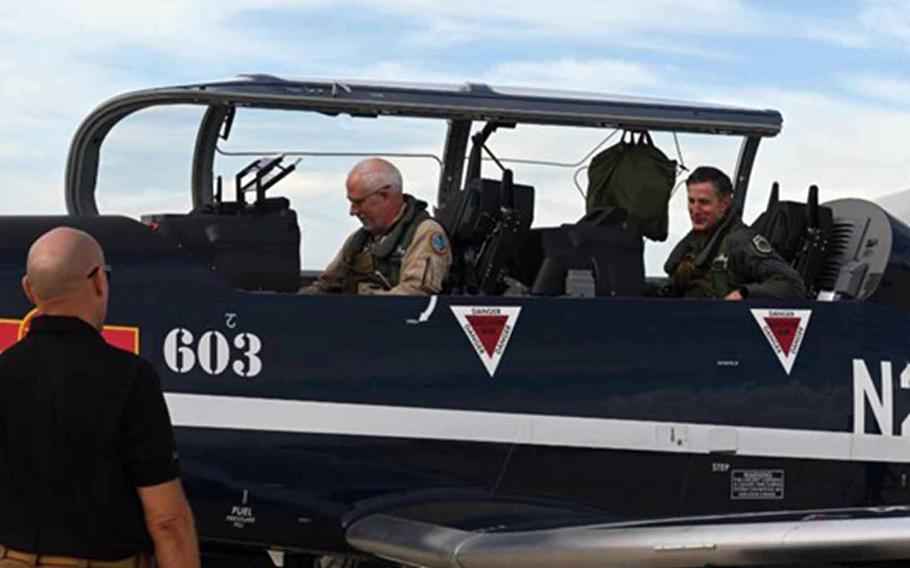 Two airmen in flight gear sit in the cockpit of a fighter jet with the canopy open.