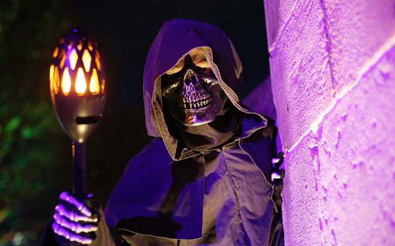 A character with the face of a skeleton wearing a cloak grasps a lit torch and peers around the corner of a stone wall at nighttime.