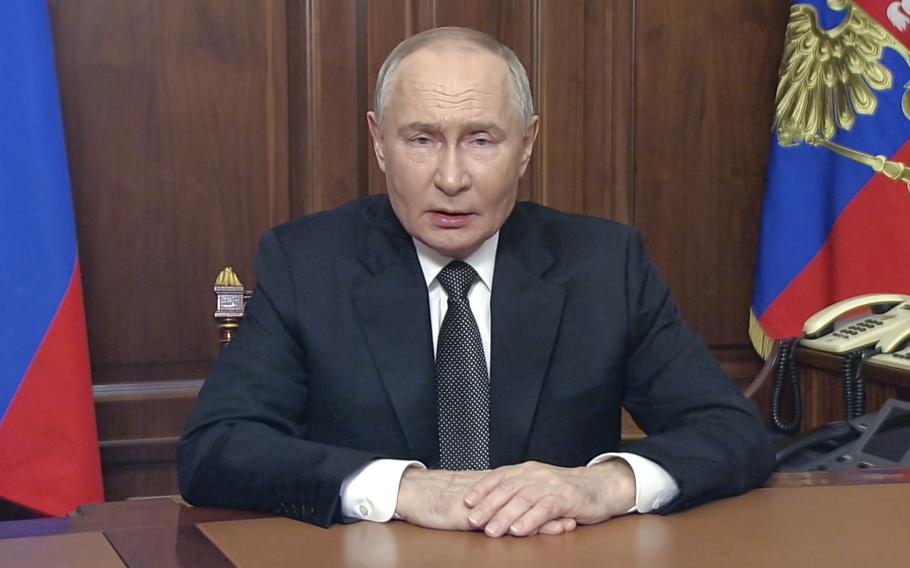 Vladimir Putin speaks while sitting at a desk in front of Russian flags.