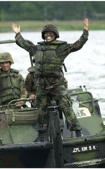 A female soldier grins and stands with her hands raised on a vehicle.