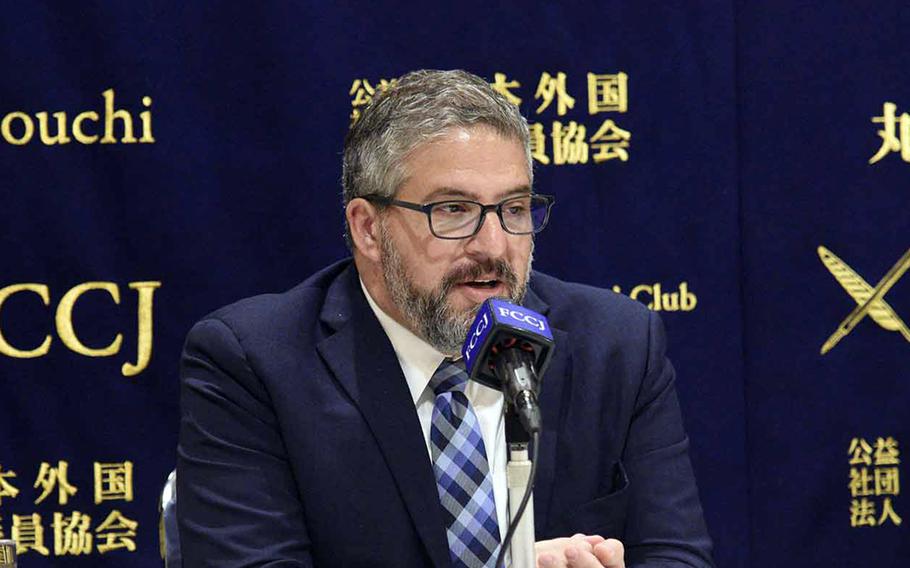 Abraham Denmark speaks into a microphone in front of a blue background.