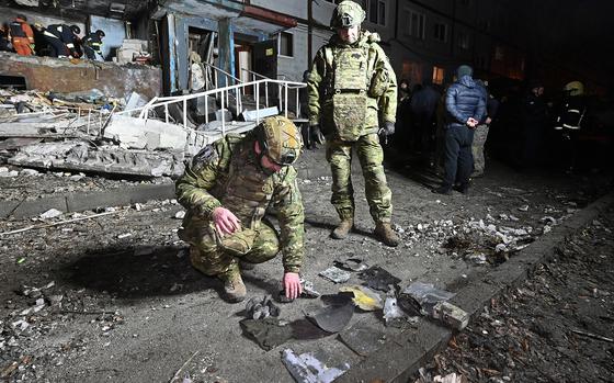 Two Ukrainian soldiers look at pieces of metal laid out on the ground outside of a damaged building.