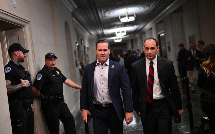 Two men in suits walk down a corrdidor, past some security or police officers.