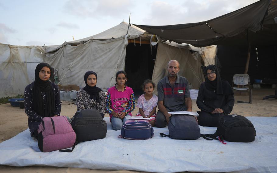 A family of six sits on the ground outside with their backpacks in front of them.