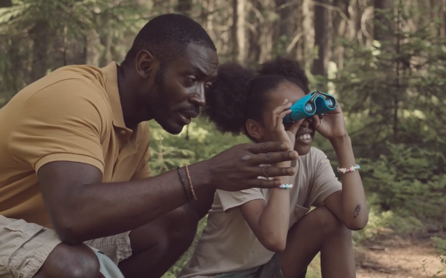 A man kneels near his child as she holds binoculars. Both are in a forest.