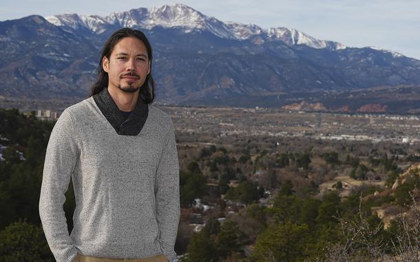 A man in a gray sweater poses in a park with a mountain landscape in the background.