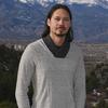 A man in a gray sweater poses in a park with a mountain landscape in the background.