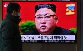 A man wearing a coat and a face mask walks past a television showing North Korean leader Kim Jong Un speaking in front of a red background.