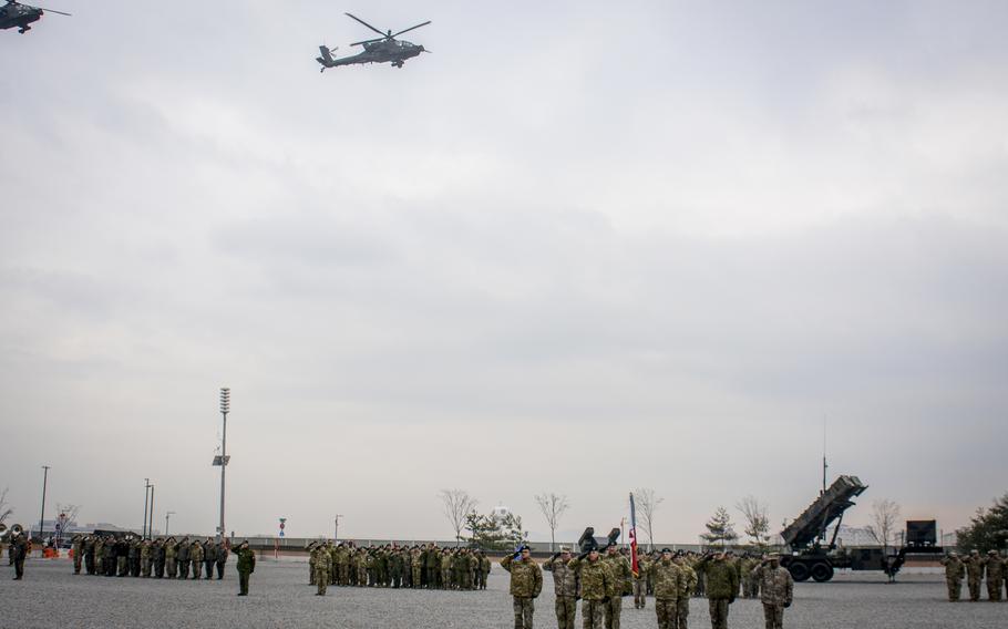 Two helicopters fly in an overcast sky above service members holding salutes.