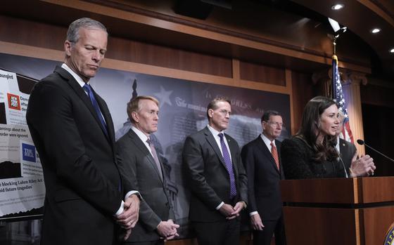 A group of men in suits stand behind a woman who speaks into a microphone at a podium.