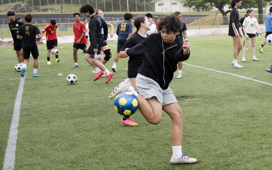 High school soccer players in warm-up jackets and shorts dribble soccer balls on a field.