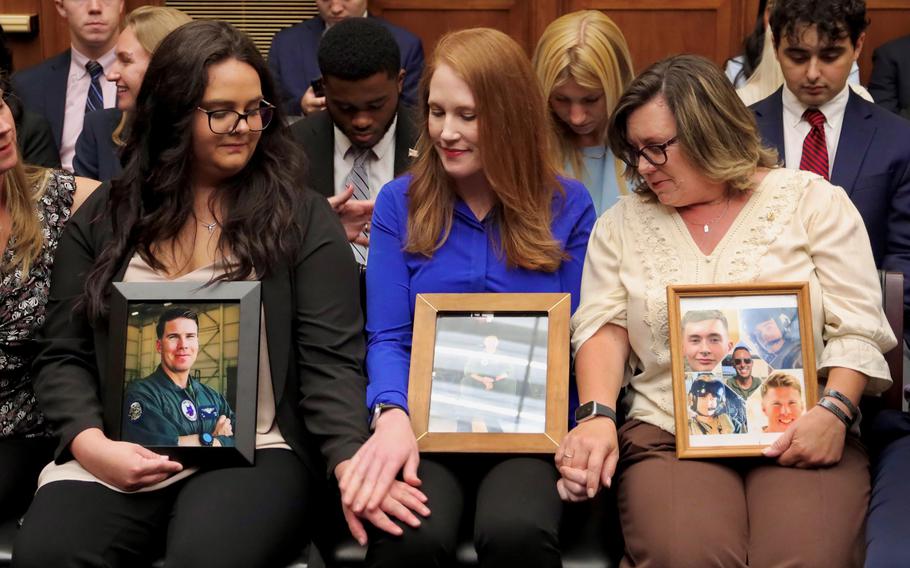 Three women sit next to each other, holding hands and framed photos of their respective loved ones who were killed in Osprey aircraft crashes.