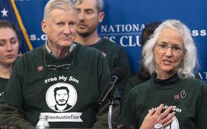 An older man and woman wear green sweatshirts with an illustration in white of their son as they speak in front of a blue wall.