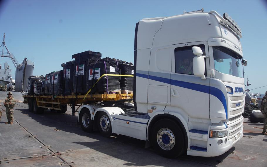 A semi-truck waits at a loading dock with its flatbed full of pallets of humanitarian aid.