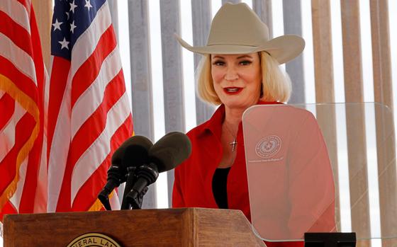 A woman wearing a cowboy hat and red jacket stands at a podium in front of a U.S. flag and speaks into a microphone.