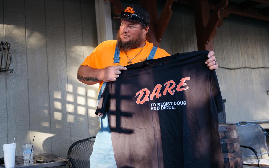 A man wearing a hat and overalls holds us a shirt that says D.A.R.E.